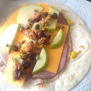 A flour tortilla wrap open with sliced meat, sliced cheddar cheese, green apple slices and veronica's health crunch.